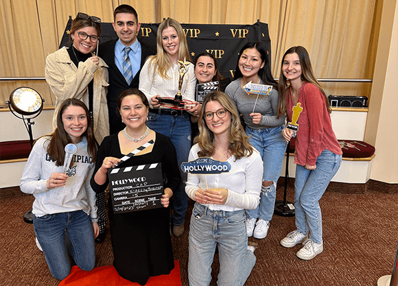 Group of nine students, eight female and one male, pose with Hollywood-themed props in front of a black VIP backdrop