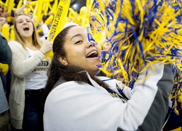 A student stands in a stadium crowd cheering and waving a pom pom