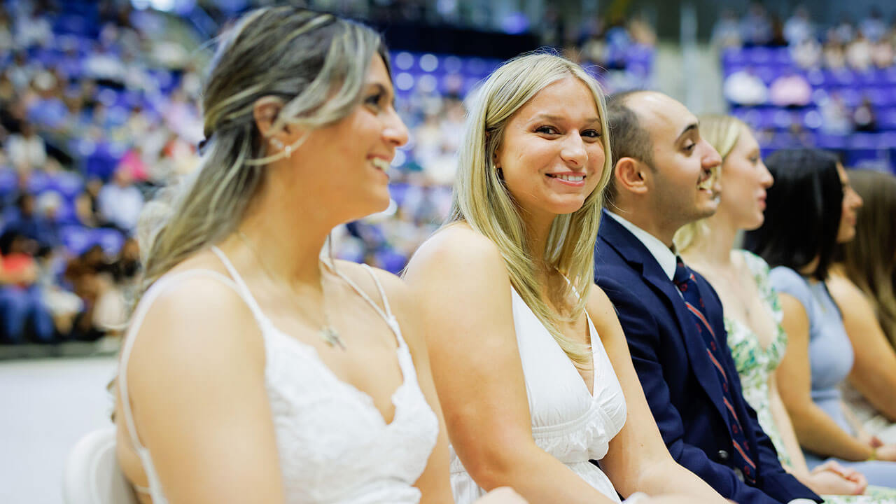 two blonde students in white dresses smile, one at the camera and the other at the presenter
