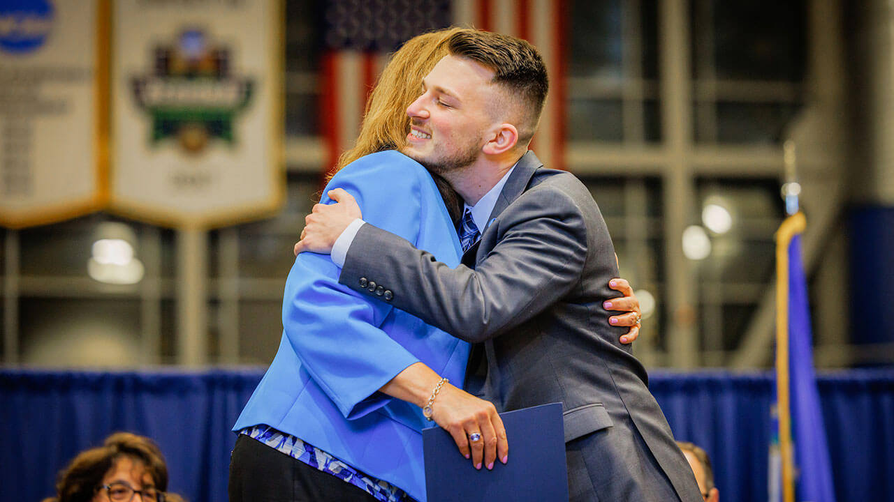 male student in gray suit hugs the school of nursing dean while being presented with his certificate