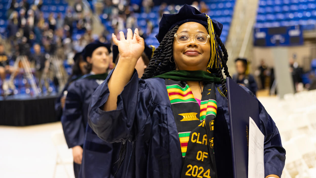 A graduate waves as she walks with her diploma under her arm