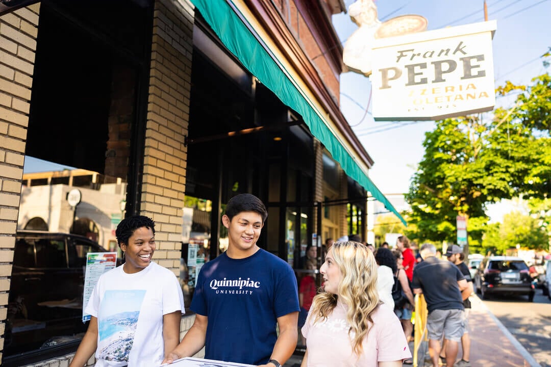 Three Quinnipiac students are walking in downtown New Haven, right behind Frank Pepe's, holding a fresh pizza.