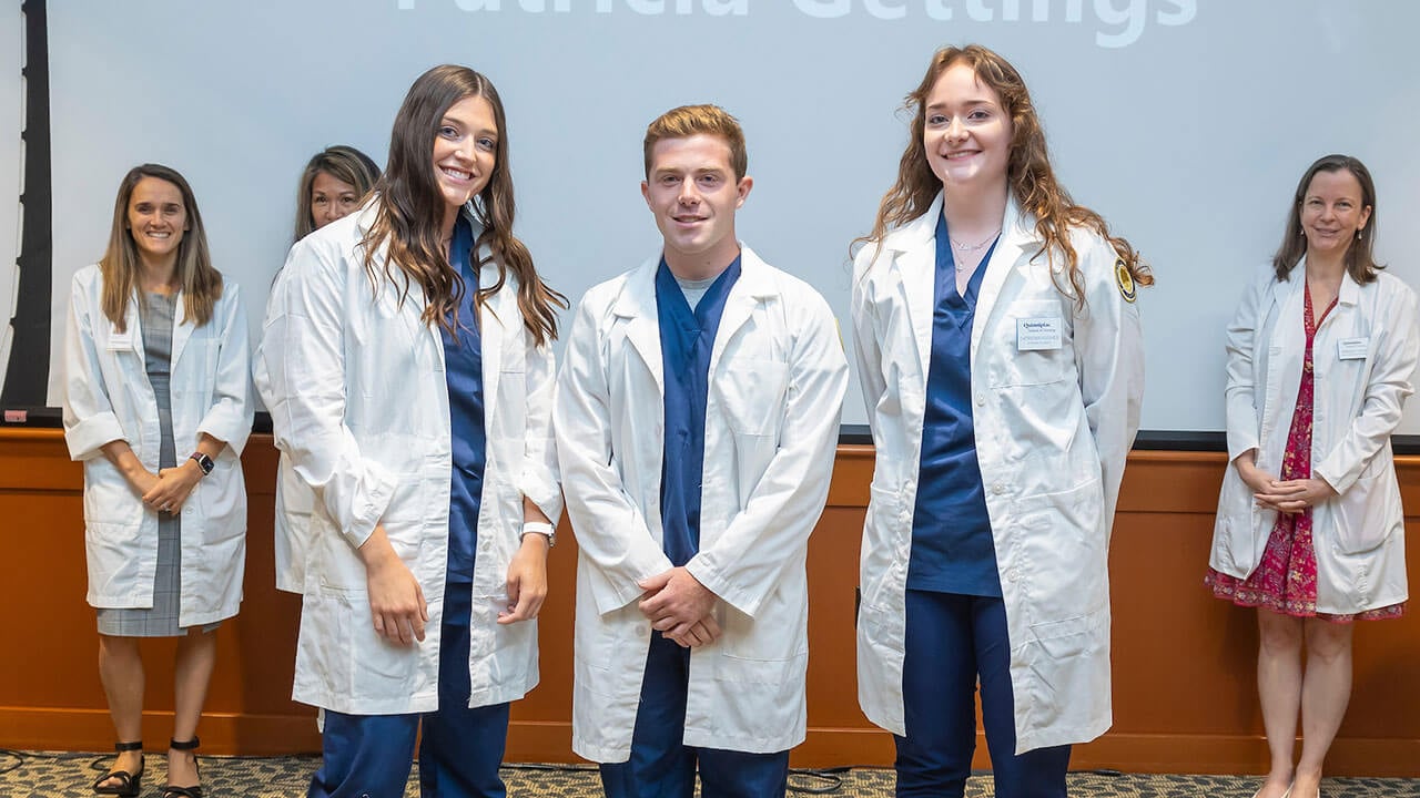 three nursing students pose in their new white coats in front of their advisors