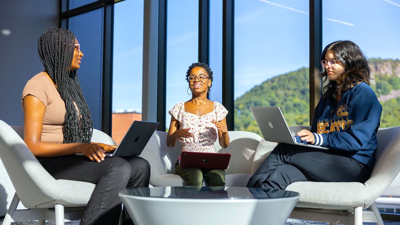 Three students sit in lounge chairs by a brightly lit window